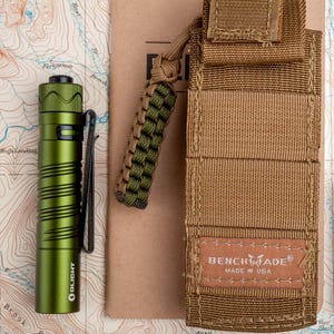 May include: A green Olight flashlight with a pocket clip and a lanyard. The flashlight is next to a brown notebook and a brown nylon pouch with the text "BENCHMADE&reg; MADE IN USA".