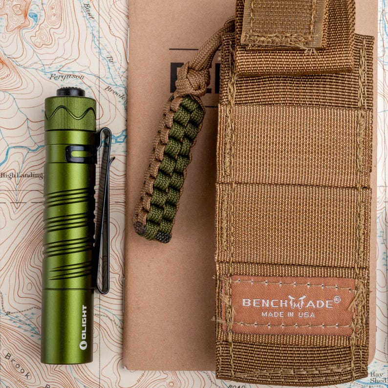 May include: A green Olight flashlight with a pocket clip and a lanyard. The flashlight is next to a brown notebook and a brown nylon pouch with the text "BENCHMADE&reg; MADE IN USA".