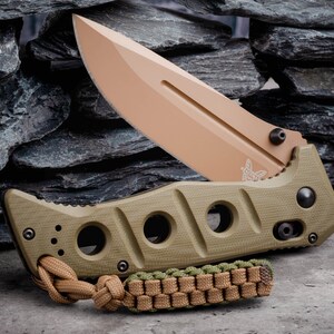 May include: A green and tan folding knife with a partially open blade. The blade is a light tan color and has a butterfly logo on it. The handle has a textured surface and a lanyard.