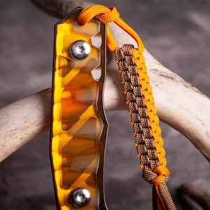 May include: Close-up of a knife with an orange and yellow handle and a braided lanyard. The handle has a textured design and is secured with two silver screws. The lanyard is brown and orange.