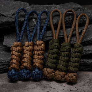 Puede incluir: Seis tiradores de cremallera de paracord dispuestos en dos grupos. El grupo izquierdo tiene tres bucles azules con cuerpos trenzados color canela y nudos azules. El grupo derecho tiene tres bucles color canela con cuerpos verde oliva.