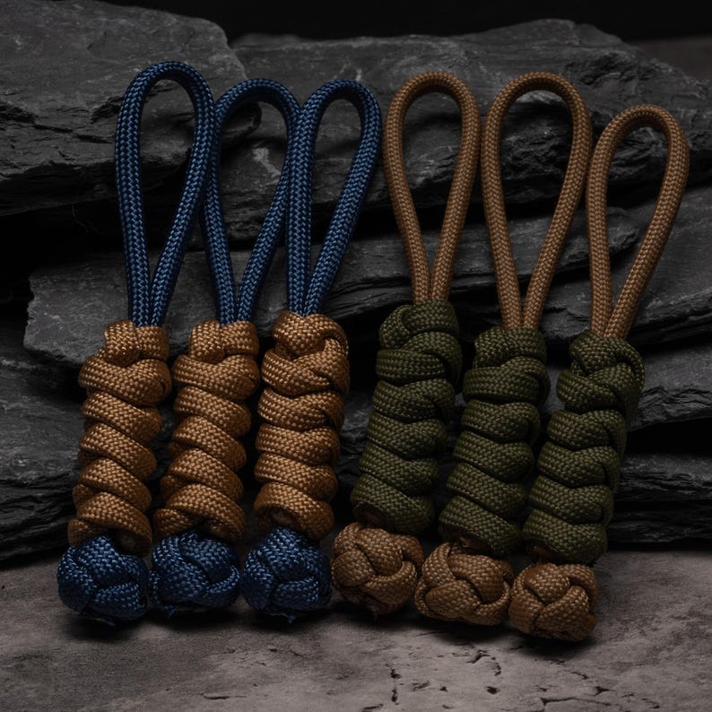 Puede incluir: Seis tiradores de cremallera de paracord dispuestos en dos grupos. El grupo izquierdo tiene tres bucles azules con cuerpos trenzados color canela y nudos azules. El grupo derecho tiene tres bucles color canela con cuerpos verde oliva.