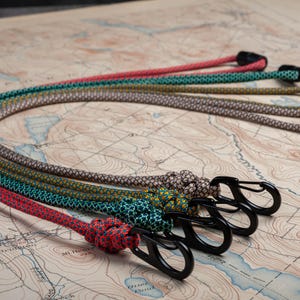 May include: Five colorful patterned keychains with black metal clips. The keychains are made of woven rope and feature different geometric patterns in shades of red, green, brown, and yellow.