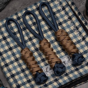 Puede incluir: Tres tiradores de cremallera hechos de paracord azul oscuro y marr&oacute;n. Cada tirador presenta un lazo en la parte superior, una secci&oacute;n central envuelta en cord&oacute;n marr&oacute;n y un nudo azul oscuro en el extremo inferior, sobre tela azul y blanca.