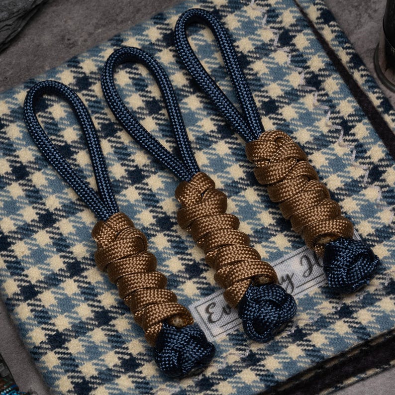 Puede incluir: Tres tiradores de cremallera hechos de paracord azul oscuro y marr&oacute;n. Cada tirador presenta un lazo en la parte superior, una secci&oacute;n central envuelta en cord&oacute;n marr&oacute;n y un nudo azul oscuro en el extremo inferior, sobre tela azul y blanca.