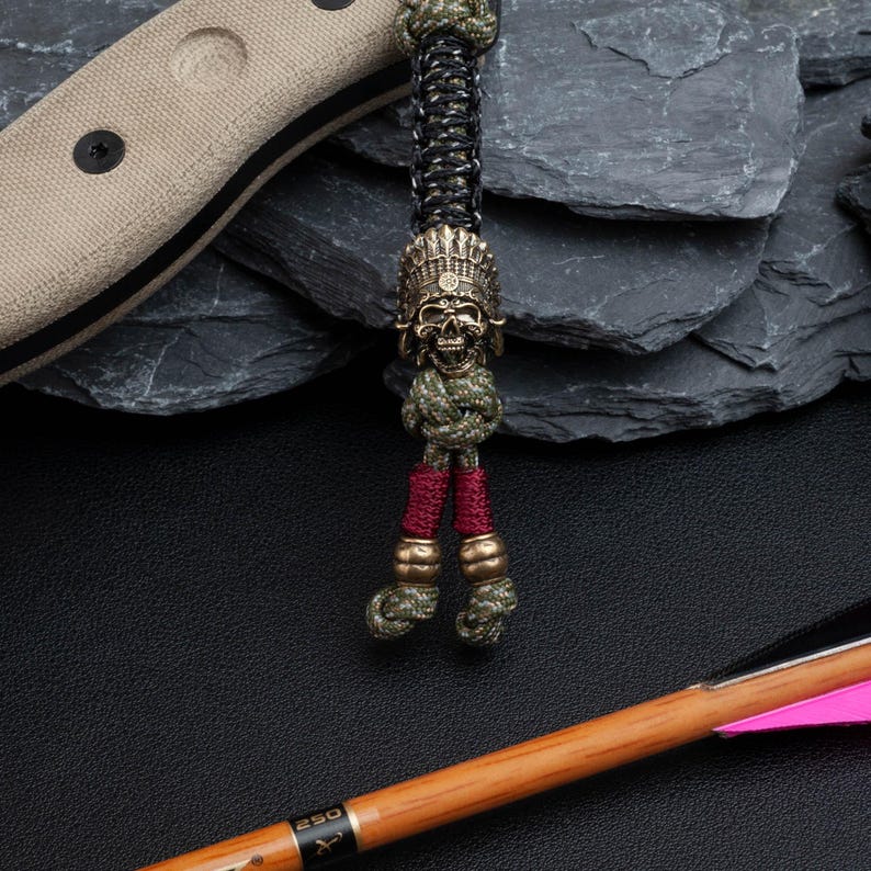 May include: A green, black, and red paracord keychain with a brass skull bead. The skull is wearing a headdress with feathers. The keychain is hanging from a knife handle.