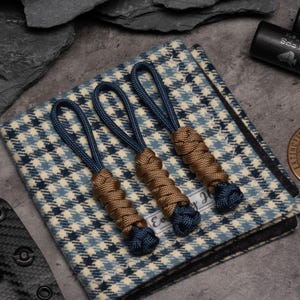 May include: Three brown and navy blue paracord keychains with a woven design. The keychains are displayed on a white and blue plaid fabric.