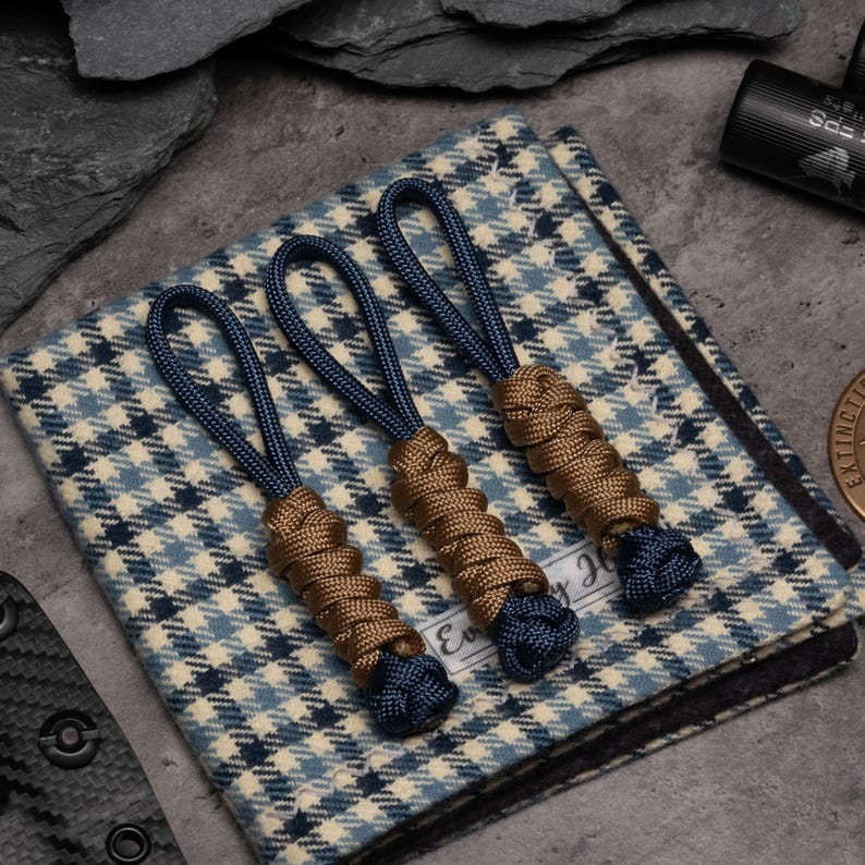 Puede incluir: Tres tiradores de cremallera con bucles de cord&oacute;n azul oscuro y cuerpos envueltos en paracord marr&oacute;n. Los tiradores est&aacute;n sobre un cuadrado de tela con patr&oacute;n de pata de gallo azul, blanco y negro con una etiqueta blanca que dice Ever.