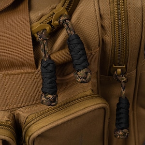 Puede incluir: Una mochila t&aacute;ctica de color marr&oacute;n claro con m&uacute;ltiples compartimentos y cremalleras resistentes. Cada tirador de cremallera est&aacute; extendido con un cord&oacute;n de paracord negro y camuflaje anudado, ofreciendo un agarre texturizado.