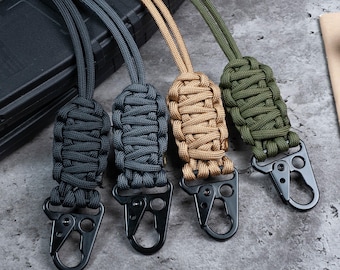 May include: Four paracord lanyards featuring intricate woven knots in black, dark gray, tan, and olive green. Each lanyard is attached to a black metal spring-loaded carabiner clip with a secure locking mechanism and a small circular cutout.