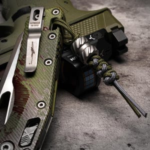 May include: Close-up of a tactical knife with a camouflage pattern and a silver pocket clip. The clip is engraved with the word "OUTBREAK" and the date "08/2025". A lanyard with a metal bead and a braided cord is attached to a small black device.