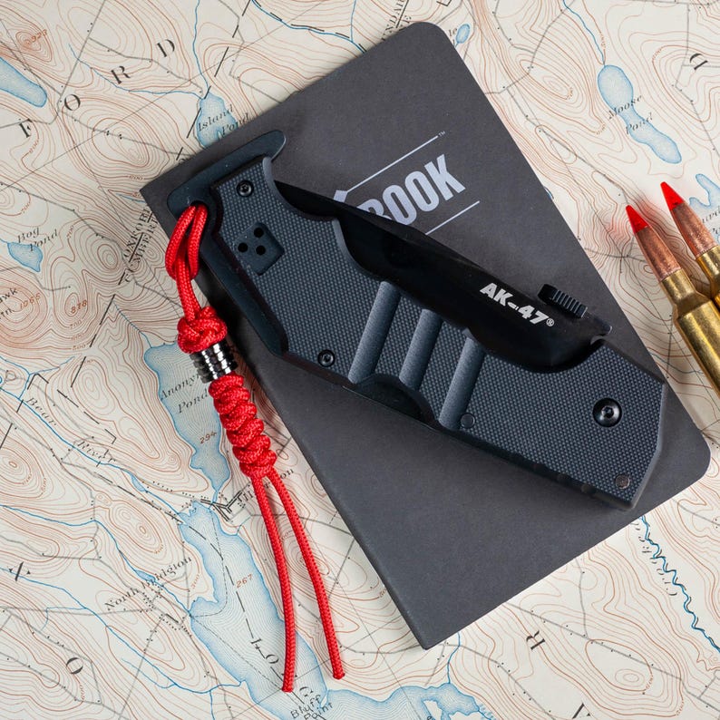 May include: A black folding knife with a red lanyard and the text "AK-47" on the blade. The knife is resting on a black notebook with the word "BOOK" on the cover. The knife is on a map.