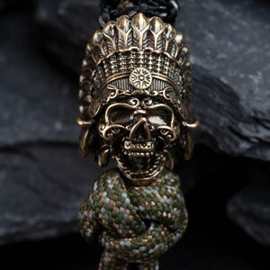May include: A gold-toned skull bead with a detailed headdress, resembling a Native American chief's headdress, is attached to a green, brown, and gray paracord.