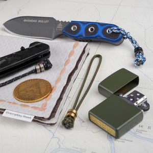 May include: An assortment of EDC (Everyday Carry) items including a knife with "Baghdad Bullet" text, a Zippo lighter, a coin, a handkerchief with "Everyday Hank" text, and a lanyard. The knife has a blue and black handle. The lighter is olive green.