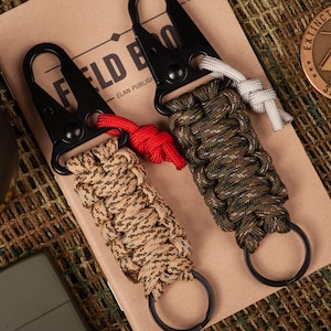 May include: Two black keychains with paracord loops. One keychain has a tan and brown camouflage pattern, and the other has a green and brown camouflage pattern. Both keychains have black metal clips and rings.
