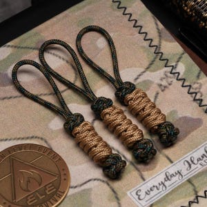May include: Three handmade zipper pulls with a loop and a braided design. The pulls feature a combination of tan, green, and black paracord. The background includes a camouflage pattern and a coin with the text "ELE".