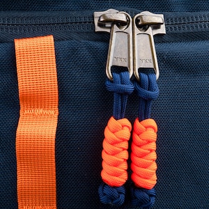 Puede incluir: Una mochila de tela azul oscuro con dos tiradores de cremallera met&aacute;licos YKK. Cada tirador est&aacute; unido a un cord&oacute;n azul oscuro con una extensi&oacute;n de nudo de paracord trenzado naranja y azul. Una correa de nailon naranja vertical a la izquierda.