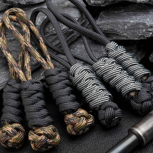 Puede incluir: Un conjunto de cinco tiradores de cremallera hechos de paracord trenzado. Dos tiradores presentan un patr&oacute;n de camuflaje con nudos negros s&oacute;lidos, mientras que tres tienen un patr&oacute;n reflectante negro y plateado con nudos negros.