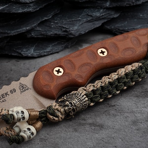 May include: A brown and tan knife with a serrated edge and a brown handle secured with gold screws. The knife has a decorative lanyard with skull beads and a bronze skull charm. The text "TEX CREEK 69" is visible on the blade.