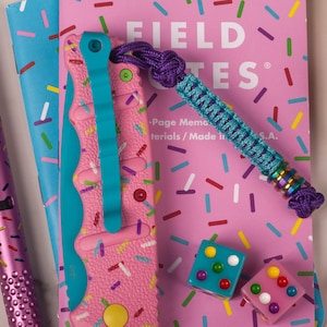 May include: A pink pocket knife with a blue handle and a sprinkle pattern. The knife is open and the blade is visible. The knife is lying on a pink notebook with the words "FIELD NOTES" printed on the cover. There are also two small dice with colorful dots on them.