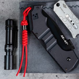 May include: A black folding knife with a red paracord lanyard and the text "AK-47" on the handle. The knife is lying on a grey fabric surface.