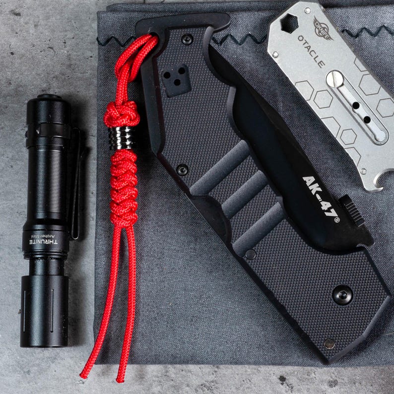 May include: A black folding knife with a red paracord lanyard and the text "AK-47" on the handle. The knife is lying on a grey fabric surface.