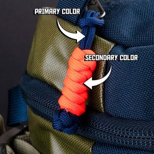 Puede incluir: Una mochila azul marino y verde oliva con un tirador de cremallera de paracord. El tirador consiste en un lazo de cord&oacute;n azul oscuro con una secci&oacute;n trenzada de color naranja brillante y un nudo azul oscuro en el extremo.