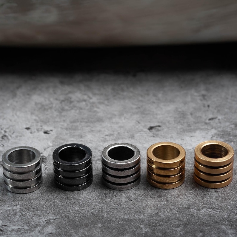 May include: Five metal rings, each with a different finish: silver, black, silver, gold, and gold. The rings are stacked in a row on a grey surface.