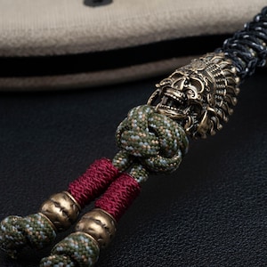 May include: A green, black, and red paracord bracelet with a brass skull bead. The skull bead has intricate details and a fierce expression.