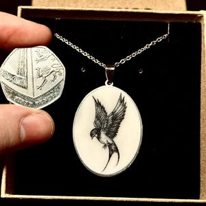 May include: Oval pendant necklace with a black and white bird design. The pendant is on a silver chain and is displayed in a gift box. A fifty pence coin is held next to the pendant for size comparison.
