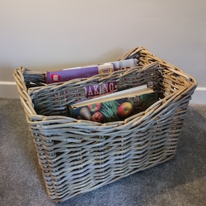 May include: A grey wicker basket containing cookbooks and magazines. The basket is rectangular with a woven pattern. The books have colourful covers, with titles including "Baking" and "Love Food".