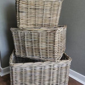 May include: Three rectangular woven wicker baskets stacked. The baskets are light grey and have a textured appearance. They are empty and suitable for storage or decoration. The baskets are made of wicker.