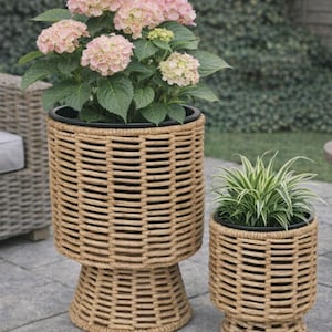 May include: Two woven wicker plant pots, one tall and one short, each containing plants. The taller pot holds a pink hydrangea with green leaves. The smaller pot has a green and white plant. Both pots are on a stone patio.