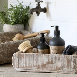 May include: A whitewashed wooden box containing two black pump bottles labelled "Eternal" and two wooden brushes. The box is sitting on a wooden surface.