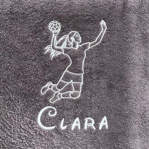 Könnte beinhalten: Ein graues Handtuch mit weißer Stickerei einer Handball spielenden Sportlerin und dem Namen "Clara" darunter.