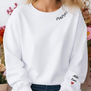 Könnte beinhalten: Ein weißes Sweatshirt mit dem Wort "Mama" und einem Herz, das auf der oberen linken Schulter gestickt ist. Der rechte Ärmel zeigt die Namen "Mila" und "Leon" mit einem roten Herz darunter. Das Sweatshirt ist ein lässiger, bequemer Stil.