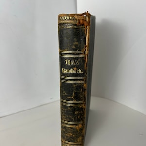 Könnte beinhalten: Ein altes, in schwarzes Leder gebundenes Buch mit goldenen Lettern auf dem Rücken, die "VEGA'S Handbuch" lesen. Das Buch ist abgenutzt und hat einige Schäden an den Kanten.