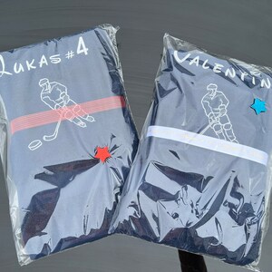 May include: Two blue hockey jerseys in clear plastic packaging. One jersey has the name "Lukas #4" printed on it and a hockey player graphic. The other jersey has the name "Valentin" printed on it and a hockey player graphic.