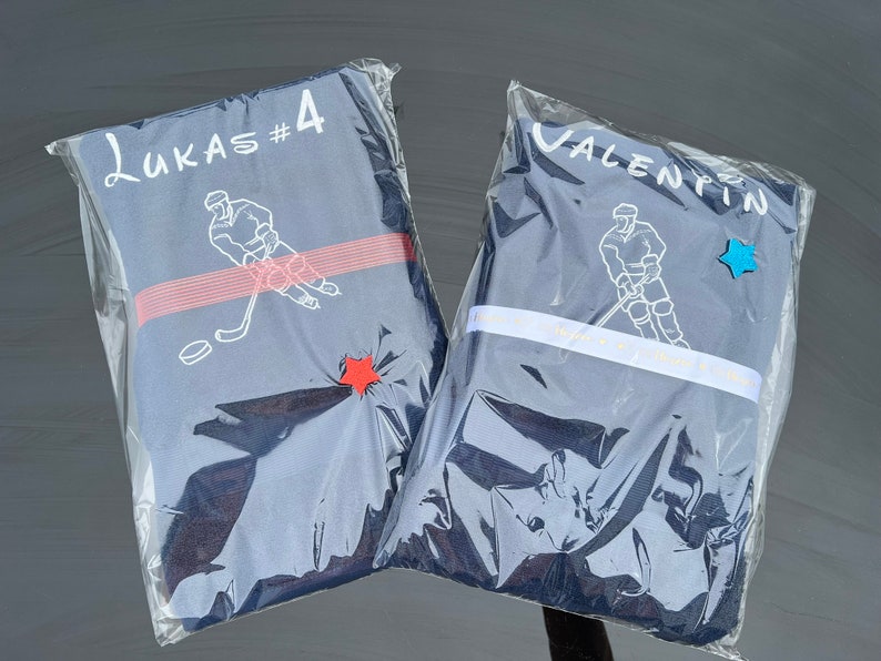 May include: Two blue hockey jerseys in clear plastic packaging. One jersey has the name "Lukas #4" printed on it and a hockey player graphic. The other jersey has the name "Valentin" printed on it and a hockey player graphic.