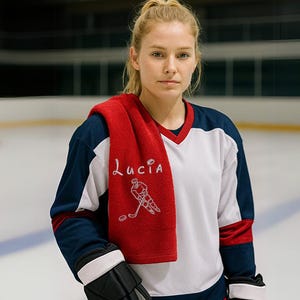 May include: A woman wearing a white and blue hockey jersey with a red towel draped over her shoulders. The towel has the name "Lucia" embroidered on it, along with a hockey player graphic. She is wearing black hockey gloves and is standing on an ice rink.