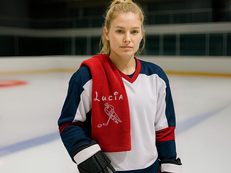 May include: A woman wearing a white and blue hockey jersey with a red towel draped over her shoulders. The towel has the name "Lucia" embroidered on it, along with a hockey player graphic. She is wearing black hockey gloves and is standing on an ice rink.