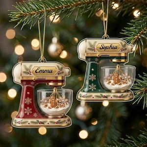 May include: Two stand mixer ornaments hanging from a tree. One is red and one is dark green, both featuring gold snowflake accents, a clear mixing bowl with a gingerbread man inside, and a nameplate reading Emma or Sophia in gold script.