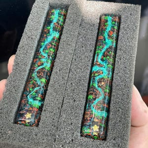 Pode incluir: Dois corpos de caneta decorativos com um padrão colorido e rodopiante de verde-azulado, laranja, verde e preto. Os corpos das canetas são exibidos em uma inserção de espuma cinza. Cada corpo de caneta tem uma estrela dourada na base.