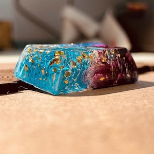 May include: A rectangular resin object with a gradient of colors. The top left is a vibrant turquoise with gold flakes, transitioning to purple and dark red on the right. The object is set against a neutral background.