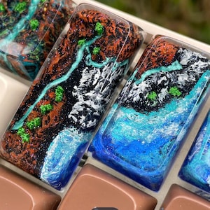 May include: Three rectangular resin keycaps featuring detailed landscape designs with textured brown mountains, bright green trees, white snowy peaks, and flowing blue rivers. The surface is glossy and clear, showcasing intricate miniature terrain.