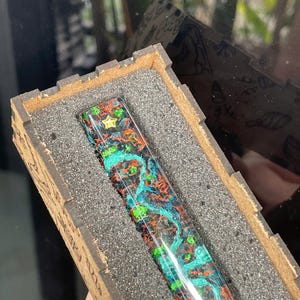 Puede incluir: Un cuerpo de bolígrafo rectangular de resina con un patrón vibrante y multicolor de pigmentos arremolinados en verde brillante, naranja y negro, acentuado por un pequeño emblema de estrella dorada incrustado cerca de la parte superior.