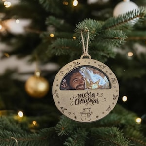 May include: A wooden Christmas ornament with a photo of a smiling person. The ornament has the words "Merry Christmas" and a snowman design. It is hanging on a Christmas tree with gold ornaments.