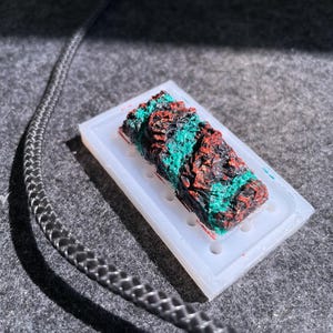 May include: A close-up of a dark brown and black resin piece with green accents. The resin piece is in a clear silicone mold.