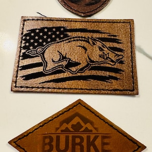 May include: Three brown leather patches with laser-engraved designs. The top shield-shaped patch shows a mountain landscape. The middle rectangular patch features a wild boar over an American flag. The bottom diamond patch reads BURKE MOUNTAIN BREW.