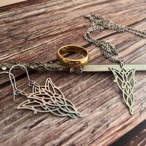 May include: A set of jewelry featuring a gold ring, a silver necklace with a pendant, and a pair of silver earrings. The pendant and earrings are in the shape of a stylized tree with intricate details.